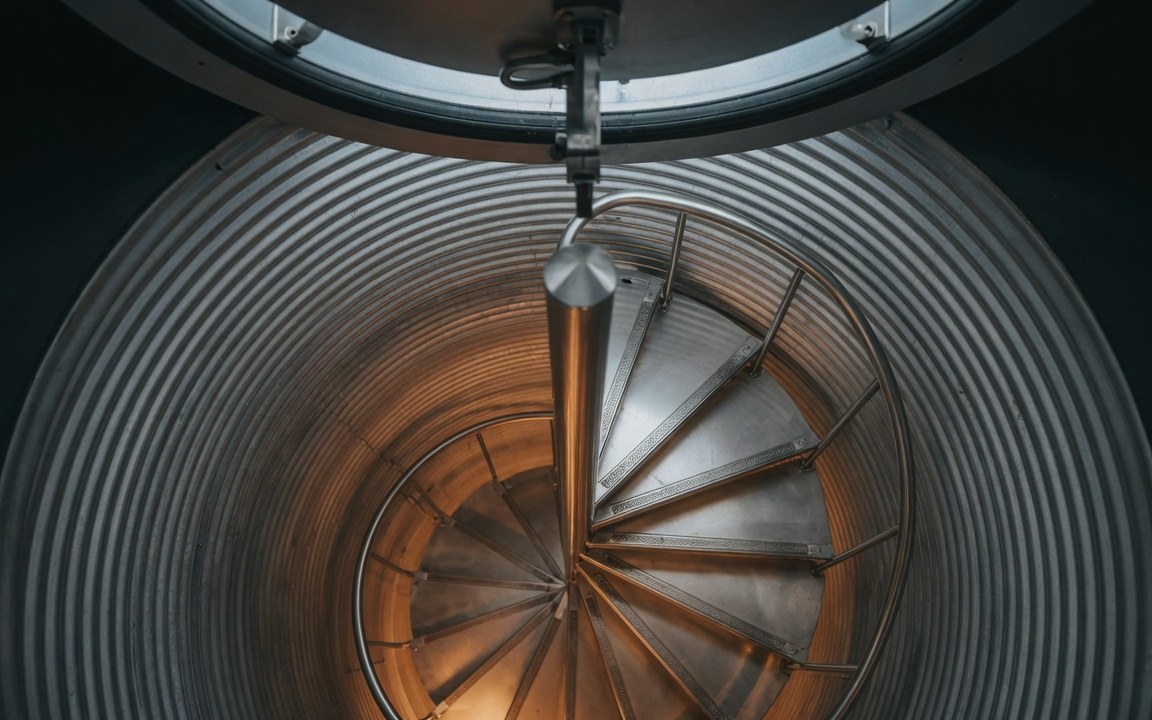 Escape tunnel — ø120 cm galvanized CMP shaft with custom 304 stainless steel spiral staircase rising four metres to Madoors blast cap, low warm key light